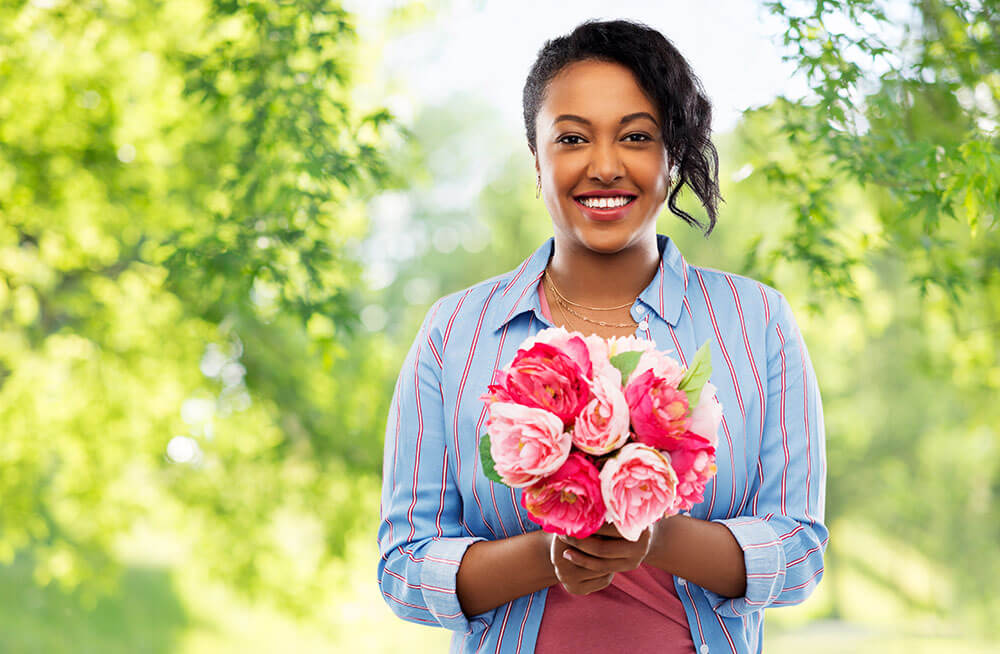 A smiling woman wearing a blue striped shirt holds a bouquet of pink and white flowers. She stands outdoors, with vibrant green trees blurred in the background, creating a bright and cheerful atmosphere.