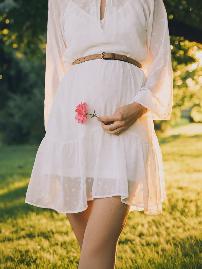 A person in a white, sheer dress holds a pink flower, standing on grass with sunlit trees in the background.