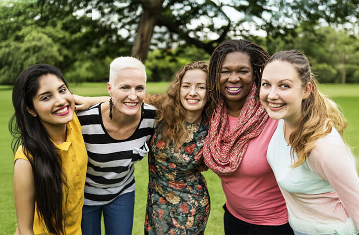 A group of five women smiling and standing close together outdoors. The scene is set in a green park with trees in the background. They are casually dressed and appear happy, enjoying each others company.